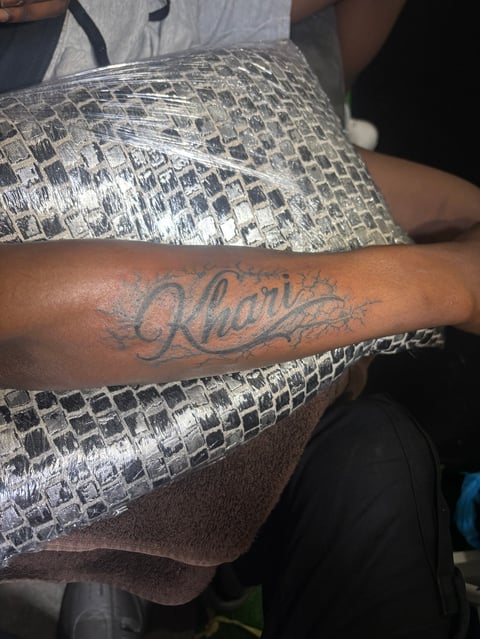 Persons arm with cursive Kharis tattoo, wrapped with a large silvery reptile or crocodile skin