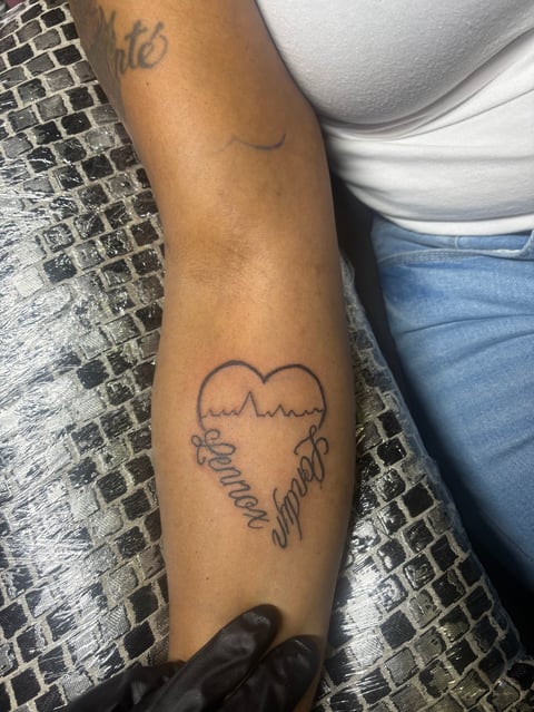 Close-up of a persons arm showing two tattoos: text at the top and a heart with wings and script text reading Somehow Yourself on the forearm