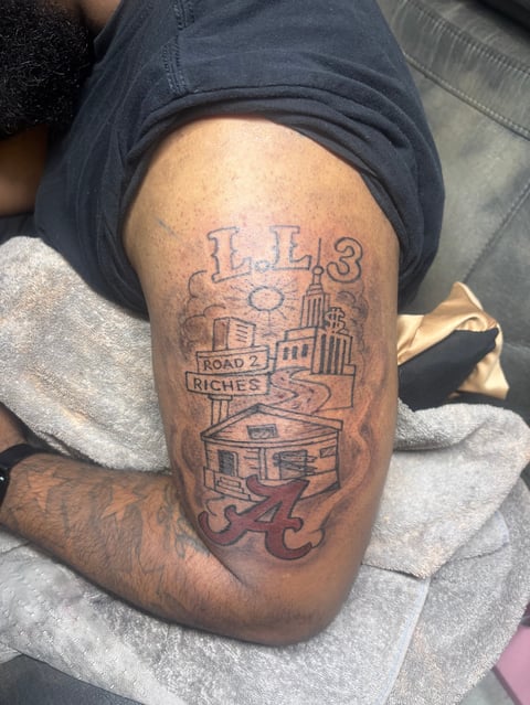 Tattoo on persons thigh featuring IL13 text with urban cityscape design and Road to Riches banner with Crimson Tide symbol