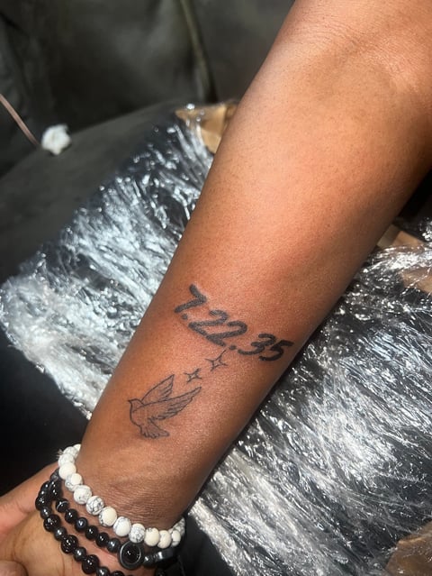 Arm with a tattoo featuring numbers and a feather design, wearing beaded bracelets, positioned over dark textured fabric