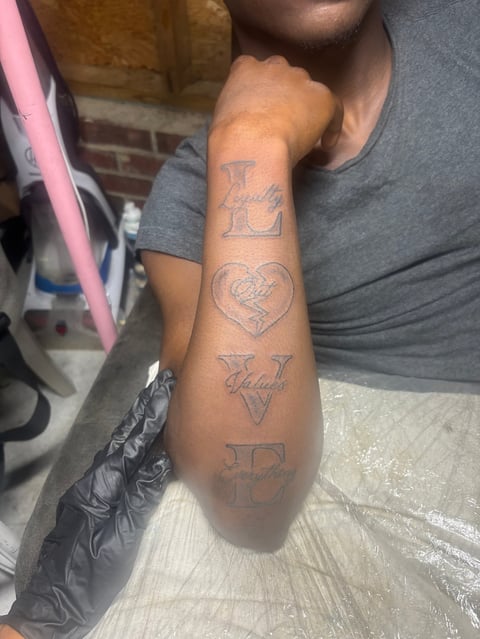 Person wearing gray shirt displaying arm tattoos including letters L and P, a heart design, and script text