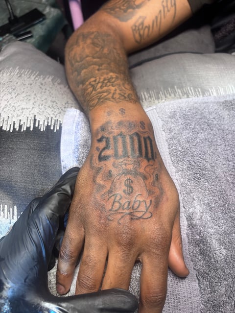 Close-up of a tattooed hand and forearm with various ink designs including script and text on brown skin
