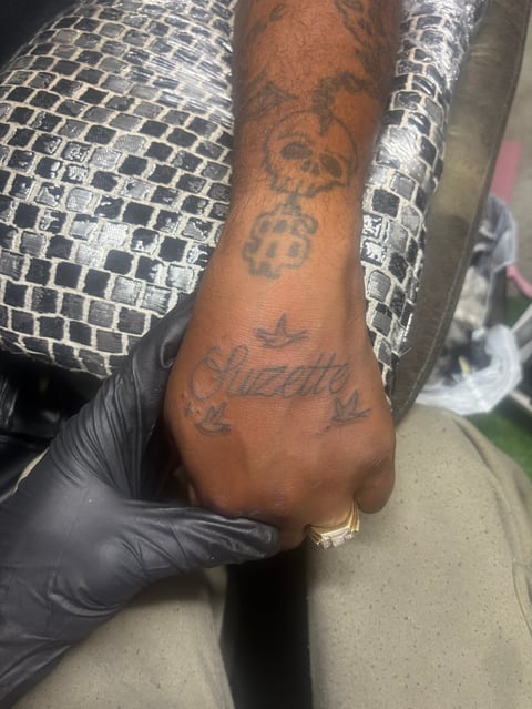 Close-up of a persons tattooed hand with script and symbol tattoos, wearing a sparkly silver jacket and black glove