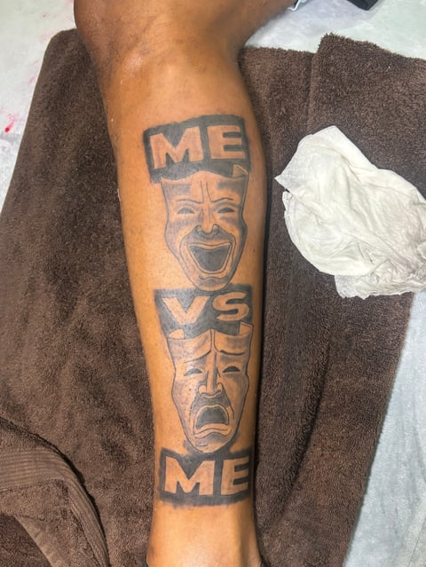 Arm tattoo featuring theatrical masks labeled ME and VS with a smiling face above and frowning face below