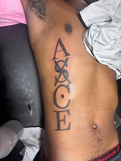 Vertical tattoo on persons ribs spelling ACE with decorative symbols between letters