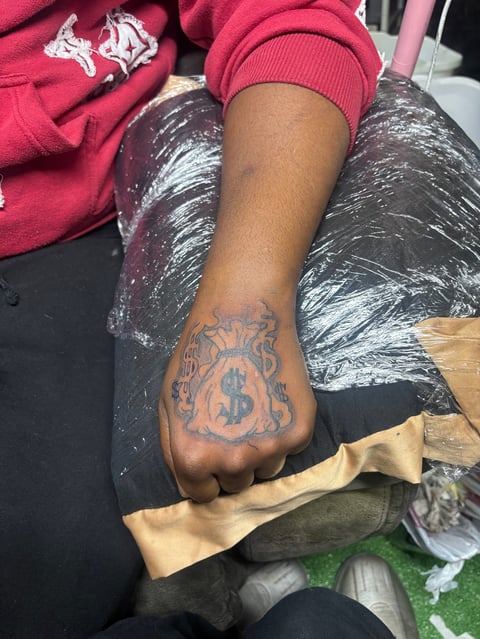 Person wearing pink hoodie displaying hand tattoo with dollar sign symbol and ornate design