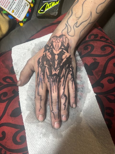 Hand tattoo of a demon or monster face with dripping ink effect on fingers, displayed on white paper with red decorative fabric in background
