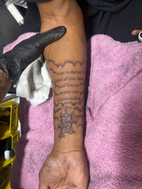 Person receiving tattoo on forearm with text and decorative design, tattoo artist wearing black gloves working on the arm