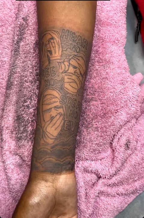 Close-up of a forearm tattoo featuring layered hands and text design on brown skin against a pink fuzzy fabric background